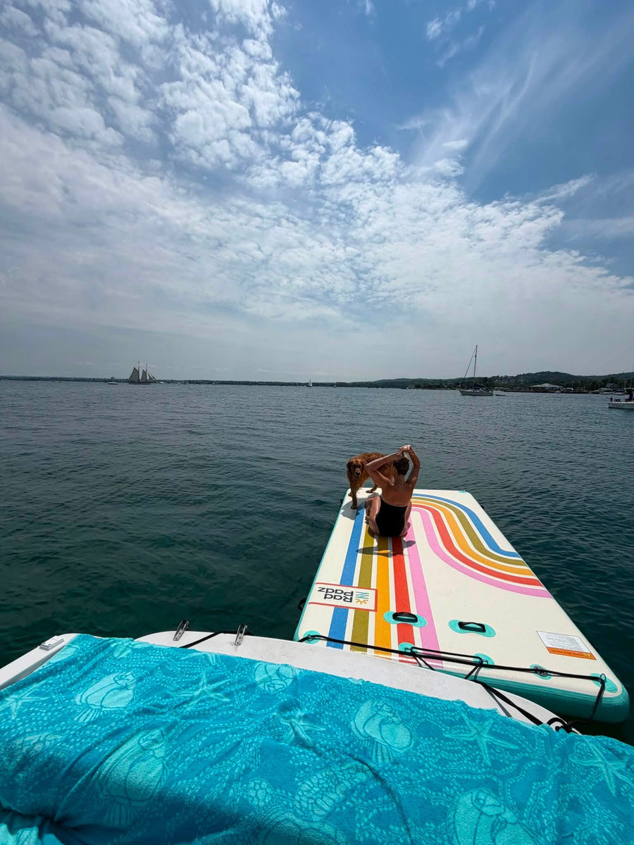 A person stands on a colorful stand-up paddleboard on the open water, with a boat deck and blue towel visible in the foreground and a cloudy sky above.